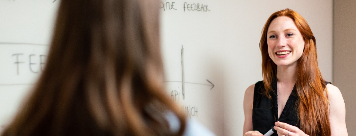 Eine Frau mit langen roten Haaren lächelt und spricht mit einer Person mit langen braunen Haaren. Sie stehen vor einem Whiteboard mit Text und einem Diagramm. Die Szene scheint ein Büro oder ein Klassenzimmer zu sein.