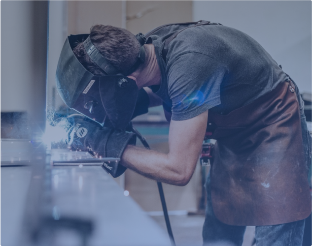 Eine Person mit Schweißhelm und Schürze beugt sich vor und schweißt aktiv Metall auf einer Werkbank. Funken sind sichtbar und der Hintergrund ist verschwommen, wodurch der Fokus auf der Schweißtätigkeit liegt.