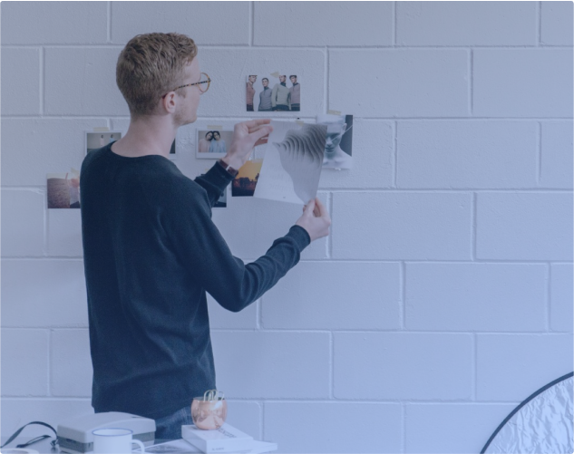 Eine Person mit Brille und schwarzem Pullover pinnt Fotos und Kunstdrucke an eine weiße Backsteinwand. Auf einem Tisch daneben liegen verschiedene Gegenstände, darunter ein kleines elektronisches Gerät und eine Kerze. Die Szene hat eine kreative und lockere Atmosphäre.
