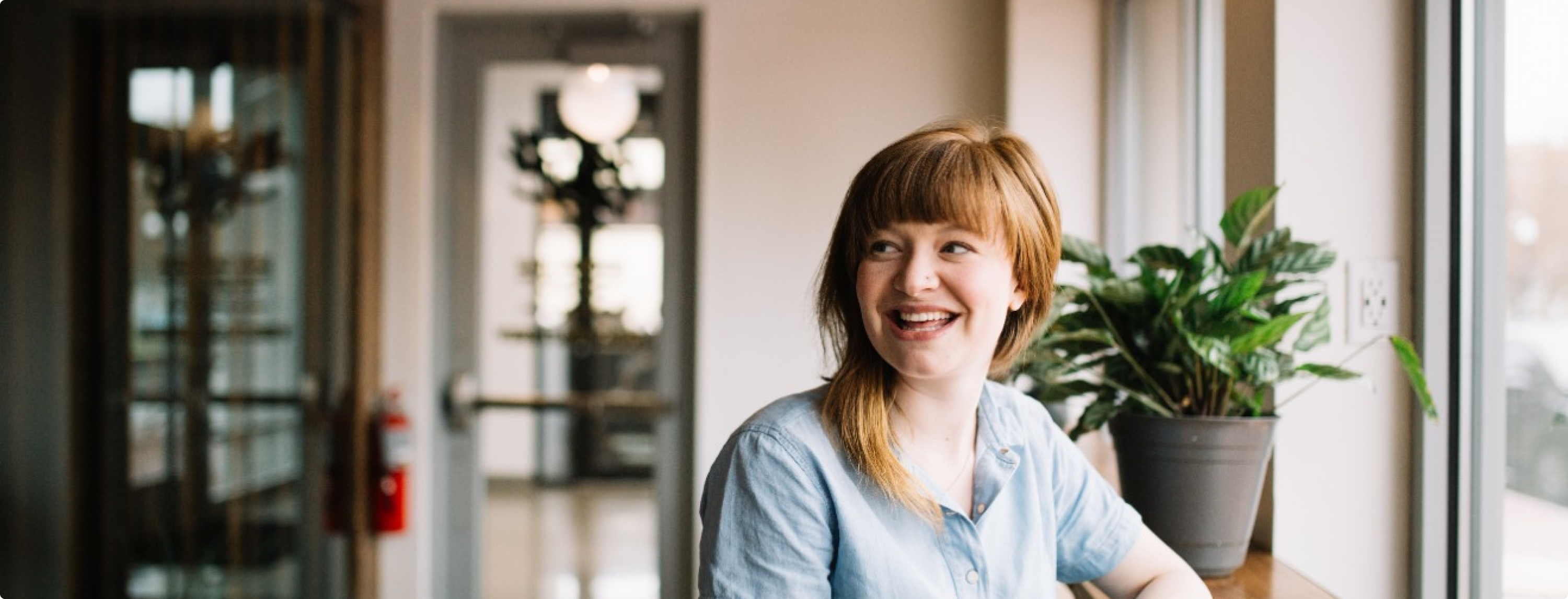 Eine Person mit langen roten Haaren sitzt lächelnd an einem hellen Fenster. Sie trägt ein hellblaues Hemd und blickt nach rechts. Auf der Fensterbank steht eine Topfpflanze, im Hintergrund ist ein Flur mit verschwommenen Details zu sehen.