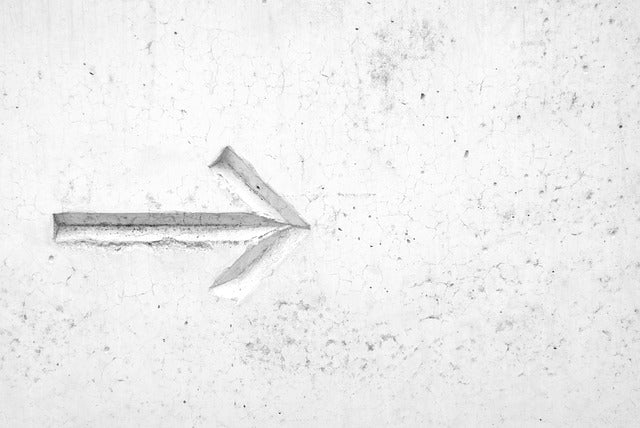 Der Aufpreis besteht aus einer strukturierten weißen Wand mit einem nach rechts zeigenden Pfeil, der in die Oberfläche eingraviert ist, um Schattenpunkte zu erzeugen. Die Wand weist Gebrauchsspuren und kleine Risse auf.