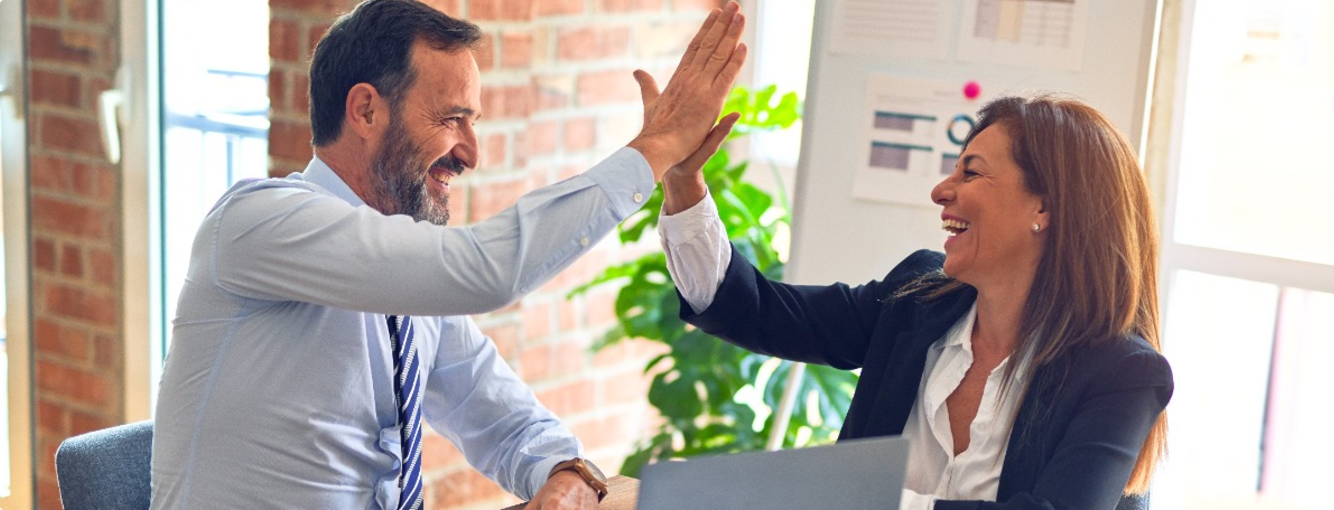 Ein Mann und eine Frau in Business-Kleidung geben sich in einem hellen Büro ein High-Five. Sie sitzen an einem Schreibtisch, vor ihnen steht ein Laptop. Im Hintergrund sind eine Pflanze und große Fenster mit Jalousien zu sehen. Beide lächeln und wirken begeistert.