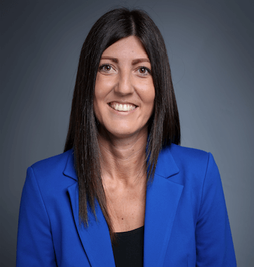 Eine Frau mit langen dunklen Haaren lächelt in die Kamera. Sie trägt einen hellblauen Blazer über einem schwarzen Oberteil. Der Hintergrund ist schlicht dunkelblau-grau und sorgt für ein professionelles Erscheinungsbild.