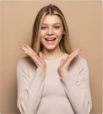 Eine lächelnde Frau mit langen blonden Haaren und einem hellbeigen Pullover streckt ihre Hände in einer freudigen Geste vor einem schlichten beigen Hintergrund hoch.