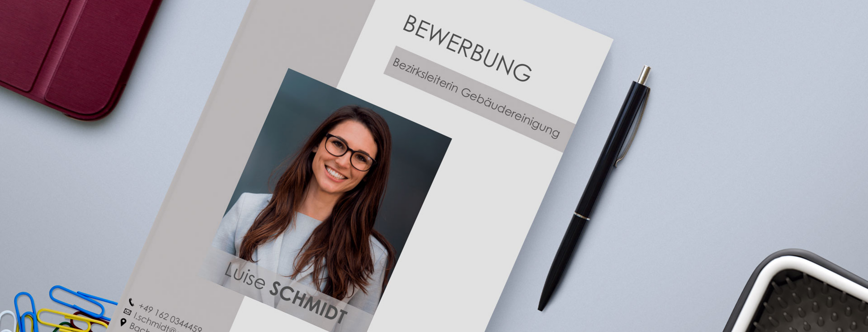 Ein Bewerbungsschreiben mit dem Titel „Bewerbung“ und einem Foto einer lächelnden Person mit Brille. Das Dokument liegt auf einem Schreibtisch neben einem Stift, Büroklammern, einem roten Notizbuch und einem Stifthalter. Kontaktdaten und Name sind teilweise sichtbar.