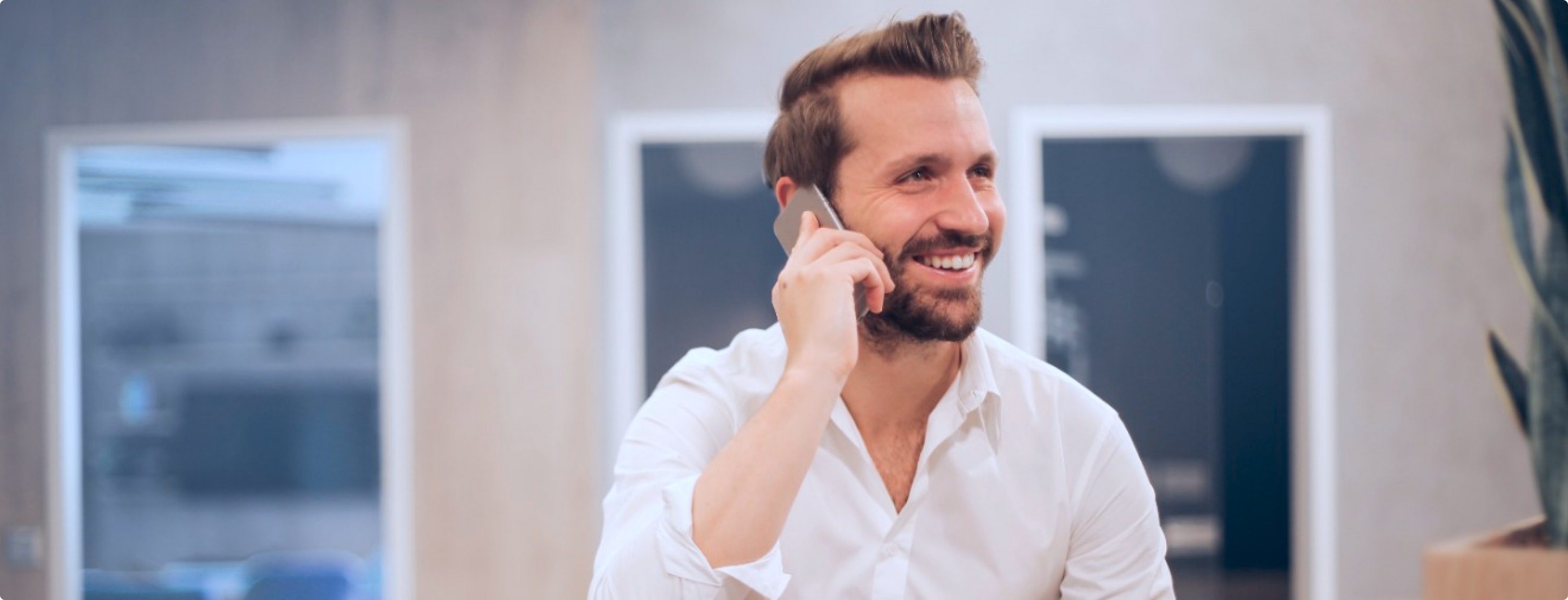 Ein Mann mit Bart und weißem Hemd lächelt, während er mit seinem Smartphone spricht. Er befindet sich in einem Gebäude, im Hintergrund sind unscharfe Bürotüren und Pflanzen zu sehen.