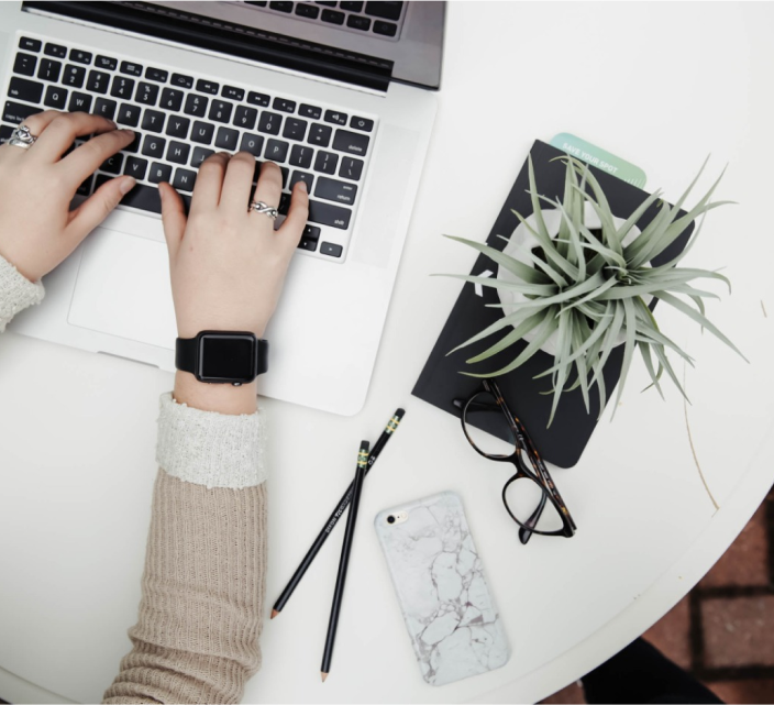 Hände tippen auf einer Laptop-Tastatur. Am Handgelenk trägt man eine Smartwatch. In der Nähe auf dem runden Tisch stehen eine kleine Pflanze, ein Notizbuch, eine Brille, zwei Bleistifte und ein Smartphone mit einer Hülle mit Marmormuster.