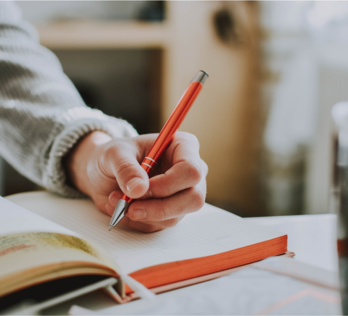 Eine Person hält einen roten Stift und schreibt in ein Notizbuch. Das Notizbuch liegt aufgeschlagen auf einem Tisch, im Vordergrund ist ein verschwommenes Buch zu sehen. Die Person trägt einen grauen Pullover. Die Atmosphäre wirkt warm und konzentriert.