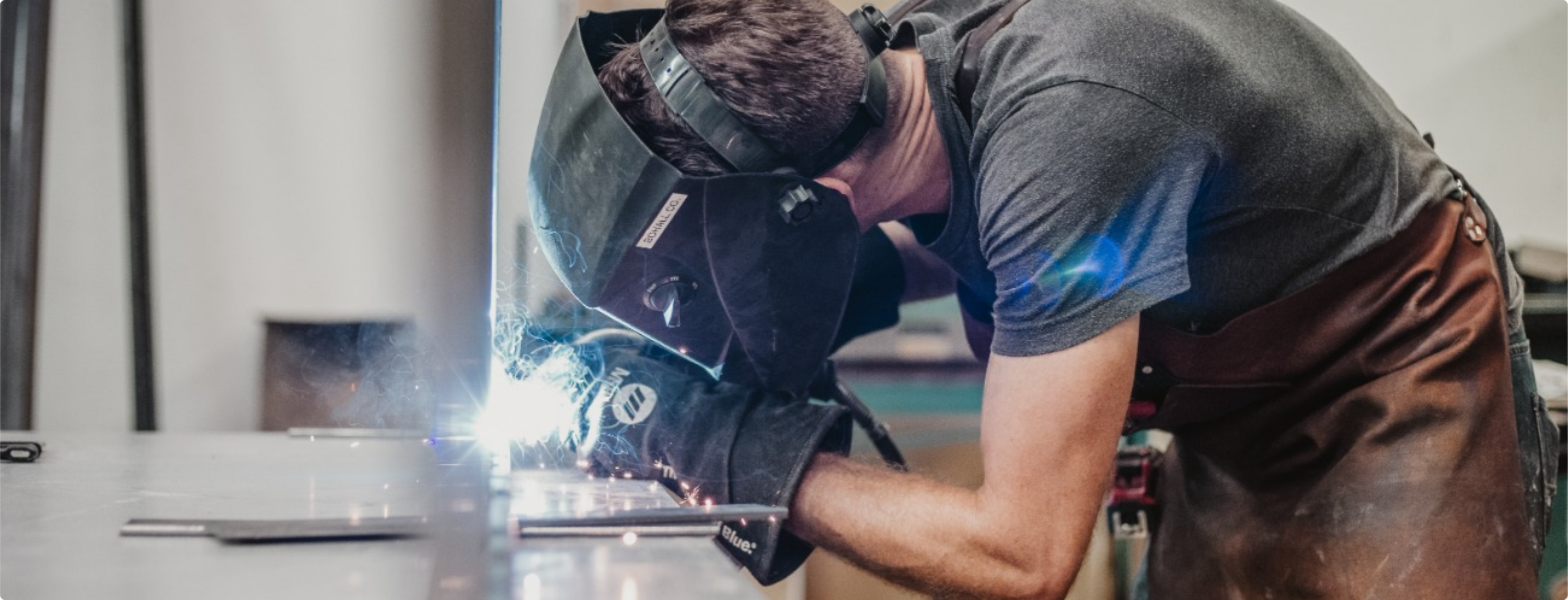 Ein Schweißer in Schutzkleidung konzentriert sich auf das Schweißen eines Metallstücks, wobei helle Funken entstehen. Er beugt sich über eine Werkbank in einer Werkstatt und trägt aus Sicherheitsgründen Handschuhe und eine Schürze.