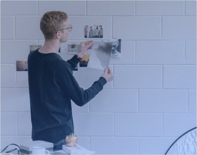 Eine Person mit schwarzem Hemd und Brille ordnet Fotos an einer weißen Backsteinwand. Sie hält ein Foto hoch, während andere bereits angepinnt sind. Auf einem Tisch in der Nähe stehen eine Kamera und andere Fotoausrüstung.