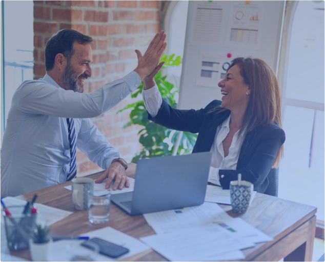 Ein Mann und eine Frau in Business-Kleidung geben sich an einem Holzschreibtisch mit Laptop, Papieren und Kaffeetassen ein High-Five. Sie lächeln beide und sitzen in einem Büro mit einer Backsteinwand und einem Fenster im Hintergrund.