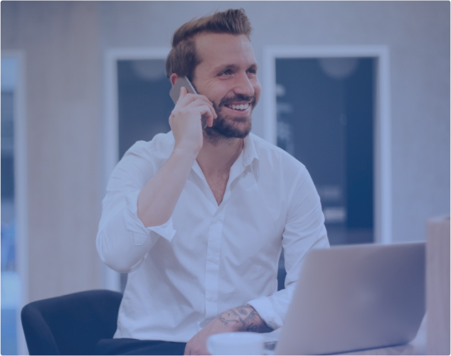 Ein lächelnder Mann mit Bart sitzt in einem Büro und telefoniert mit seinem Smartphone. Er trägt ein weißes Hemd und hat einen Laptop vor sich. Im Hintergrund ist eine moderne Büroumgebung mit verschwommenen Details zu sehen.