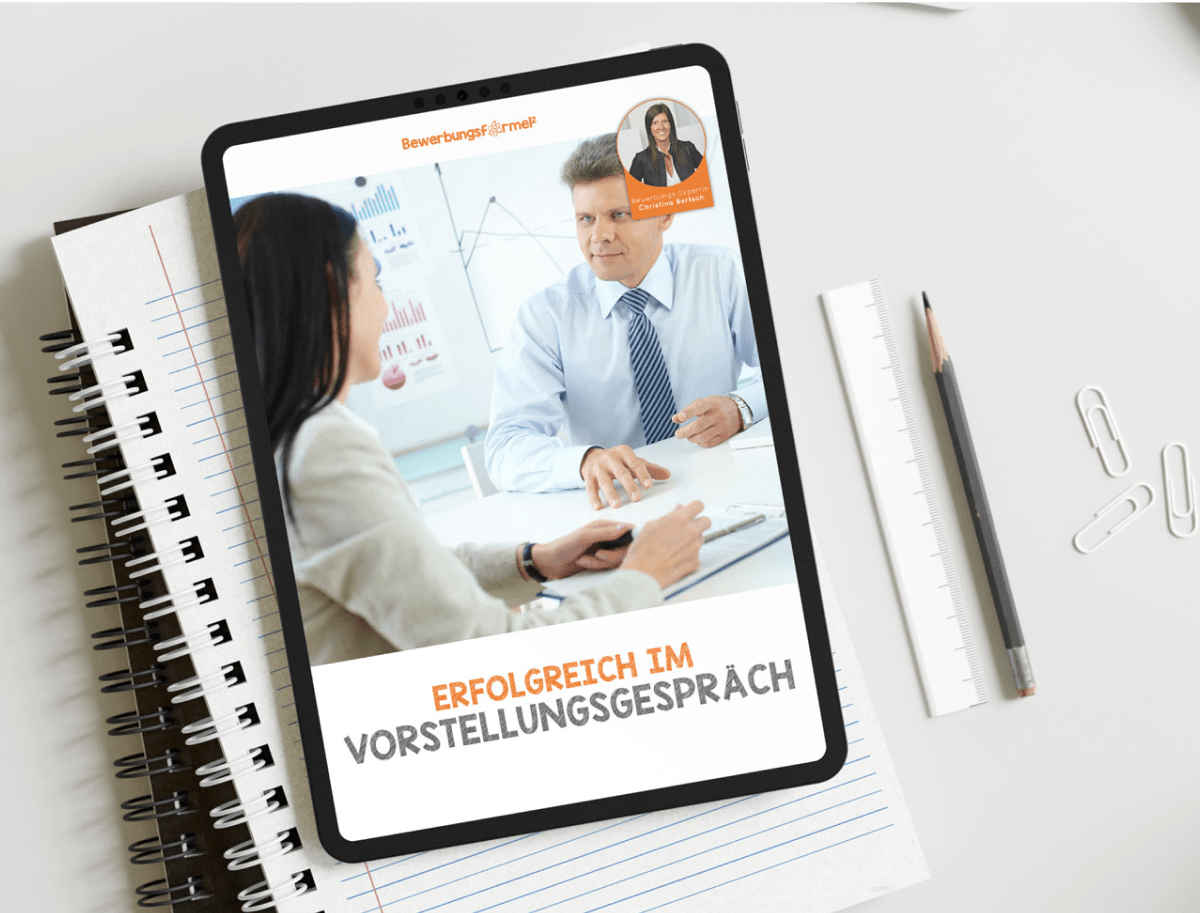 Auf einem Tablet wird ein E-Book mit dem Titel „Erfolgreich im Vorstellungsgespräch (PDF)“ angezeigt, das einen Mann und eine Frau in einer beruflichen Umgebung zeigt. Es ruht auf einem Spiralblock, umgeben von einem Bleistift, Büroklammern und Notizen einer Bewerbungsexpertin zum Umgang mit kniffligen Fragen im Vorstellungsgespräch.