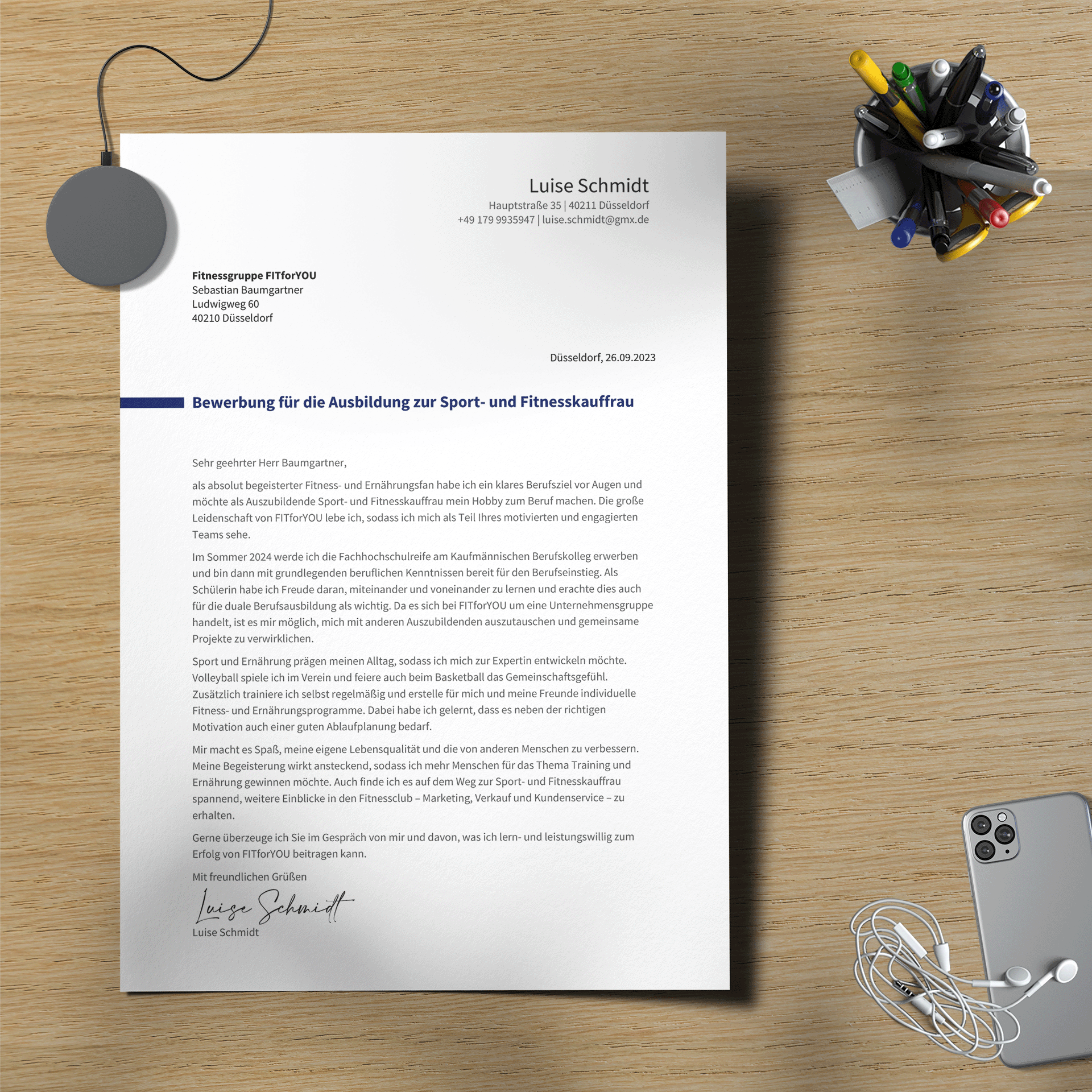 Ein „Bewerbungsschreiben Nachwuchstalente“ liegt auf einem Holzschreibtisch, umgeben von Büroutensilien wie Büroklammern, einem Stifteköcher mit bunten Markern, Kopfhörern und einem Smartphone. Der Brief ist an ein Fitnessunternehmen adressiert und von Luise Schmidt unterschrieben.